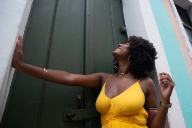 Afro saçlı, sarı elbiseli, yeşil kapıya dayanmış fotoğraf için poz veren güzel siyahi kadın portresi. Yolculukta mutlu insan.