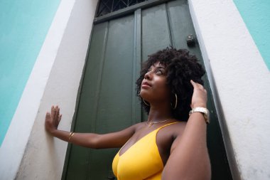 Afro saçlı, sarı elbiseli, yeşil kapıya dayanmış fotoğraf için poz veren güzel siyahi kadın portresi. Yolculukta mutlu insan.