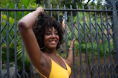 Eski demir bir çite dayanan sarı bir elbise giyen afro saçlı güzel siyah bir kadının portresi. Yolculuktaki mutlu insan..