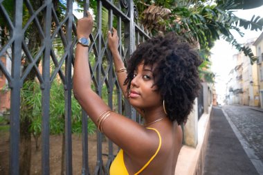 Eski demir bir çite dayanan sarı bir elbise giyen afro saçlı güzel siyah bir kadının portresi. Yolculuktaki mutlu insan..