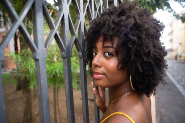 Eski demir bir çite dayanan sarı bir elbise giyen afro saçlı güzel siyah bir kadının portresi. Yolculuktaki mutlu insan..