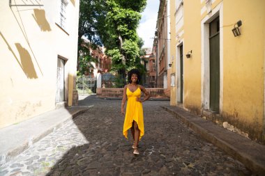 Güzel siyah bir kadın kaldırım taşı sokaklarda yürüyor sarı giysiler içinde, afro saçlı, gülümseyen ve mutlu. Yolculukta kendine güveni olan biri.