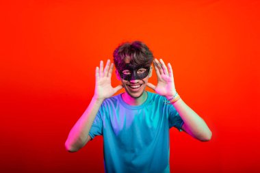 Karnaval maskesi takan yakışıklı genç adam kameraya bakıp el hareketi yapıyor. Turuncu neon arka planda izole edilmiş. Brezilya