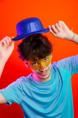 Mavi gömlekli, karnaval maskesi takan ve elinde mavi şapka tutan mutlu ve eğlenceli bir genç. Kameraya poz veriyorum. Neon turuncu arka planda izole edilmiş. Parti için hazır..