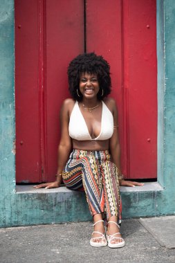 Güzel siyah güçlü kadın eski kırmızı bir kapının önünde oturuyor. Gülümseyen ve güçlü bir insan. Brezilya