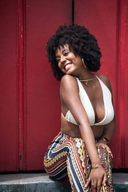 Güzel siyah güçlü kadın eski kırmızı bir kapının önünde oturuyor. Gülümseyen ve güçlü bir insan. Brezilya