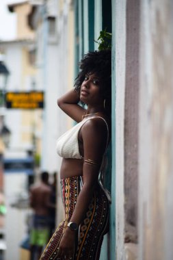 Arkası duvara dönük, fotoğraf için poz veren güzel siyahi bir kadının yarım boy fotoğrafı. Gülümseyen ve güçlü bir insan. Brezilya