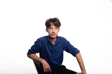 Siyah saçlı, mavi gömlekli, oturan ve kameraya ciddi ciddi bakan genç ve yakışıklı bir öğrenci. Beyaz arka planda izole edilmiş. Kendine güvenen biri. Profesyonel konsept.