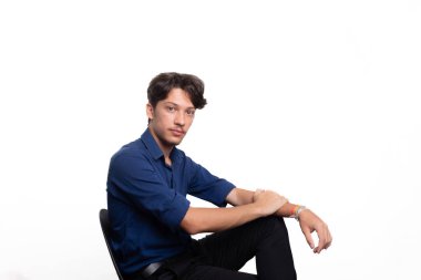 Siyah saçlı, mavi gömlekli, oturan ve kameraya ciddi ciddi bakan genç ve yakışıklı bir öğrenci. Beyaz arka planda izole edilmiş. Kendine güvenen biri. Profesyonel konsept.