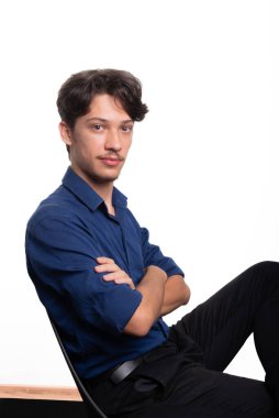 Siyah saçlı, mavi gömlekli, oturan ve kameraya ciddi ciddi bakan genç ve yakışıklı bir öğrenci. Beyaz arka planda izole edilmiş. Kendine güvenen biri. Profesyonel konsept.