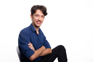 Siyah saçlı, mavi gömlekli, oturan, gülümseyen ve kameraya bakan yakışıklı bir genç. Beyaz arka planda izole edilmiş. Kendine güvenen biri. Profesyonel konsept.