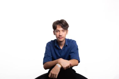 Siyah saçlı, mavi gömlekli, oturan ve kameraya ciddi ciddi bakan genç ve yakışıklı bir öğrenci. Beyaz arka planda izole edilmiş. Kendine güvenen biri. Profesyonel konsept.