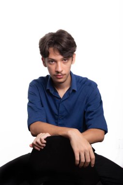 Siyah saçlı, mavi gömlekli, oturan ve kameraya ciddi ciddi bakan genç ve yakışıklı bir öğrenci. Beyaz arka planda izole edilmiş. Kendine güvenen biri. Profesyonel konsept.
