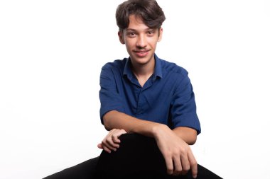 Siyah saçlı, mavi gömlekli, oturan ve kameraya ciddi ciddi bakan genç ve yakışıklı bir öğrenci. Beyaz arka planda izole edilmiş. Kendine güvenen biri. Profesyonel konsept.