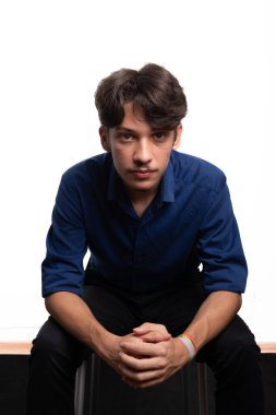 Siyah saçlı, mavi gömlekli, oturan ve kameraya ciddi ciddi bakan genç ve yakışıklı bir öğrenci. Beyaz arka planda izole edilmiş. Kendine güvenen biri. Profesyonel konsept.