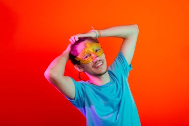 Karnaval maskesi takan mutlu ve eğlenceli genç adam iki eliyle saçına dokunuyor. Neon turuncu arka planda izole edilmiş. Bahia Karnavalı.