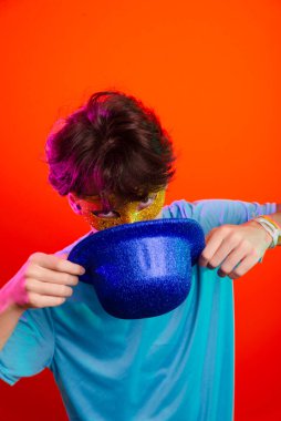 Mavi gömlekli, karnaval maskesi takan ve elinde mavi şapka tutan mutlu ve eğlenceli bir genç. Kameraya poz veriyorum. Neon turuncu arka planda izole edilmiş. Parti için hazır..