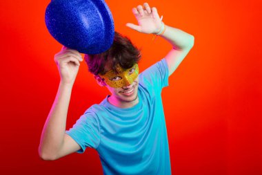 Mavi gömlekli, karnaval maskesi takan ve elinde mavi şapka tutan genç bir adam. Kameraya poz veriyorum. Neon turuncu arka planda izole edilmiş. Mutlu ve eğlenceli biri..