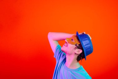 Mavi gömlekli, karnaval maskeli ve mavi şapkalı genç yukarı bakıyor. Neon turuncu arka planda izole edilmiş. Mutlu ve eğlenceli biri..