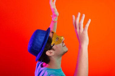 Mavi gömlekli, karnaval maskeli ve mavi şapkalı genç yukarı bakıyor. Neon turuncu arka planda izole edilmiş. Mutlu ve eğlenceli biri..