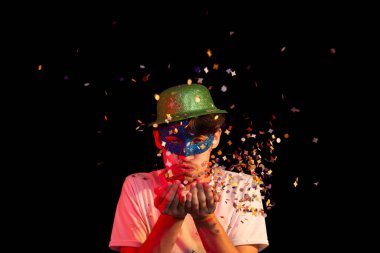 Beyaz gömlekli, karnaval maskesi takan ve yeşil şapkalı kağıt konfetiyle eğlenen genç bir adam. Siyah arka planda izole edilmiş. Mutlu ve neşeli kişi.