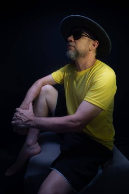 Olgun bir adam, 50 'li yaşlarda, sarı gömlek, güneş gözlüğü ve şapka takıyor, oturuyor, fotoğraf için poz veriyor. Sakin biri. Brezilya