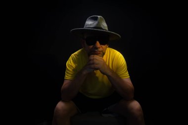 Olgun bir adam, 50 'li yaşlarda, sarı gömlek, güneş gözlüğü ve şapka takıyor, oturuyor, fotoğraf için poz veriyor. Sakin biri. Brezilya