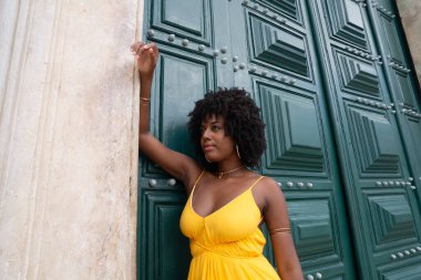 Sarı elbiseli, afro saçlı güzel siyah bir kadın büyük yeşil bir kapıya yaslanmış bir fotoğraf için poz veriyor. Mutlu ve gülümsüyor