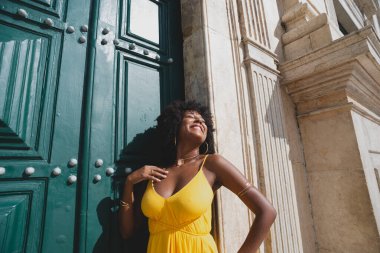 Siyah saçlı, sarı elbiseli güzel bir zenci kadın büyük yeşil kilise kapısına poz veriyor. Mutlu ve gülümsüyor