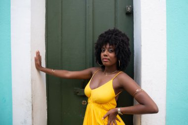 Afro saçlı, sarı elbiseli, yeşil kapıya dayanmış fotoğraf için poz veren güzel siyahi kadın portresi. Yolculukta mutlu insan.