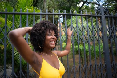 Eski demir bir çite dayanan sarı bir elbise giyen afro saçlı güzel siyah bir kadının portresi. Yolculuktaki mutlu insan..