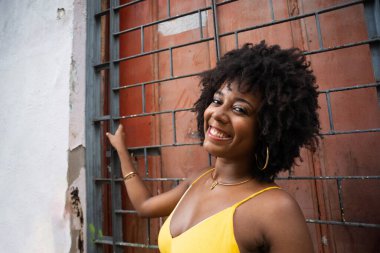 Sarı elbiseli, afro saçlı, gülümseyen demir bir çitin yanında bir kadının yakın plan portresi. Kendinden emin ve mutlu biri.