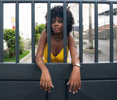 Sarı elbiseli, afro saçlı, gülümseyen demir bir çitin arkasında bir kadının portresi. Kendinden emin ve mutlu biri.