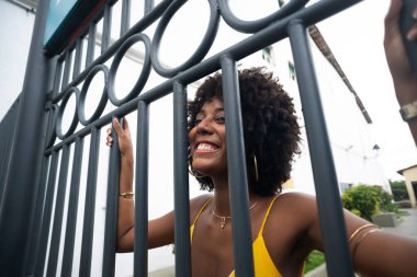 Sarı elbiseli, afro saçlı, gülümseyen demir bir çitin arkasında bir kadının portresi. Kendinden emin ve mutlu biri.