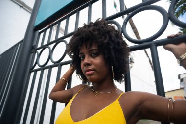 Demir bir çitin yanında sarı elbiseli, afro saçlı bir kadın. Kendinden emin ve mutlu biri.