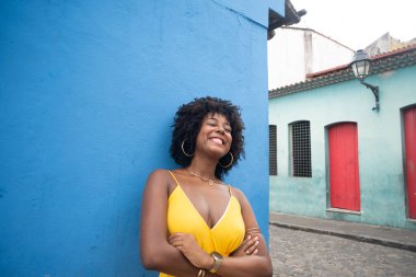 Sarı elbiseli, afro saçlı, mavi duvarda izole edilmiş ve arka planda evleri olan güzel bir kadın. Kendinden emin ve mutlu biri.