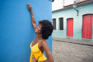 Sarı elbiseli, afro saçlı, mavi duvarda izole edilmiş ve arka planda evleri olan güzel bir kadın. Kendinden emin ve mutlu biri.