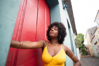 Sarı elbiseli, afro saçlı güzel bir kadın kırmızı kapının yanında fotoğraf çektirmek için poz veriyor. Kendinden emin ve mutlu biri. Brezilya