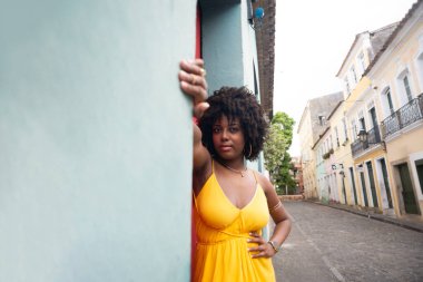 Sarı elbiseli, afro saçlı, eli cyan duvarda olan ve kameraya bakan güzel bir kadın. Kendinden emin ve mutlu biri. Brezilya