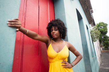 Sarı elbiseli, afro saçlı güzel bir kadın kırmızı kapının yanında fotoğraf çektirmek için poz veriyor. Kendinden emin ve mutlu biri. Brezilya