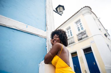 Sarı elbiseli ve afro saçlı güzel siyah kadın eski mavi bir eve yaslanıyor. Kendinden emin ve mutlu biri. Brezilya