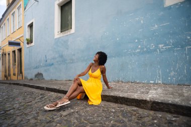 Sarı elbiseli ve afro saçlı güzel bir kadının portresi mavi duvara karşı eski caddenin kaldırımında oturuyor. Kendinden emin ve mutlu biri. Brezilya