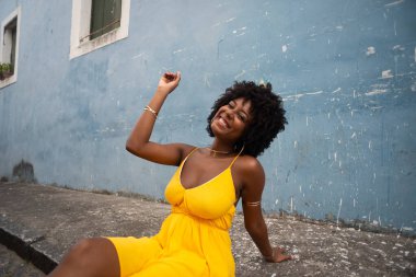 Sarı elbiseli ve afro saçlı güzel bir kadının portresi mavi duvara karşı eski caddenin kaldırımında oturuyor. Kendinden emin ve mutlu biri. Brezilya