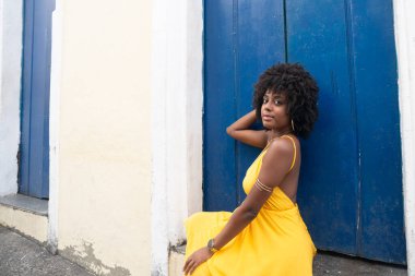 Afro saçlı, sarı elbiseli, eski bir evin mavi kapısında oturan güzel bir genç kadın. Kendine güvenen biri. Brezilya