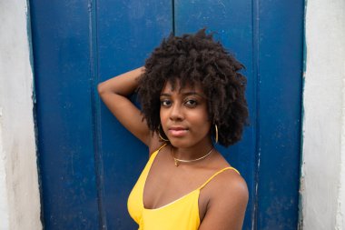 Siyah saçlı, sarı elbiseli, eski bir evin mavi kapısında oturan ve kameraya bakan genç bir kadının yakın plan portresi. Kendine güvenen biri. Brezilya