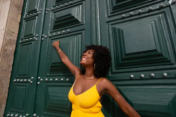 Sarı elbiseli, afro saçlı güzel siyah bir kadın büyük yeşil bir kapıya yaslanmış bir fotoğraf için poz veriyor. Mutlu ve gülümsüyor