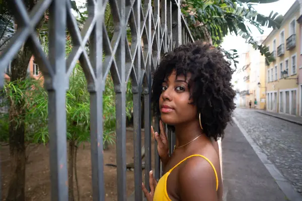 Eski demir bir çite dayanan sarı bir elbise giyen afro saçlı güzel siyah bir kadının portresi. Yolculuktaki mutlu insan..