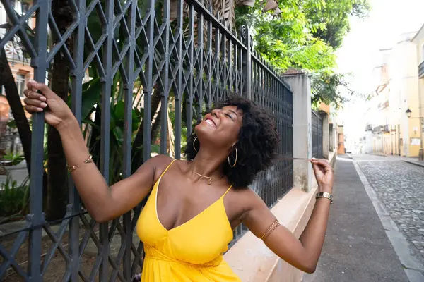 Afro saçlı, sarı elbiseli, mutlu bir kadın eski demir bir çite yaslanmış kameraya poz veriyor. Yolculuktaki mutlu insan..