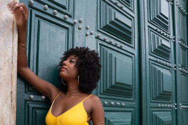 Sarı elbiseli, afro saçlı güzel siyah bir kadın büyük yeşil bir kapıya yaslanmış bir fotoğraf için poz veriyor. Mutlu ve gülümsüyor