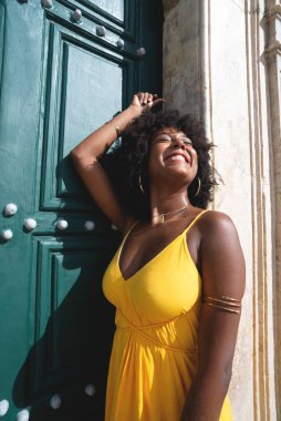 Siyah saçlı, sarı elbiseli güzel bir zenci kadın büyük yeşil kilise kapısına poz veriyor. Mutlu ve gülümsüyor